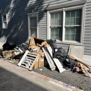 A large pile of construction debris, cardboard boxes, and black trash bags piled against the side of a house.